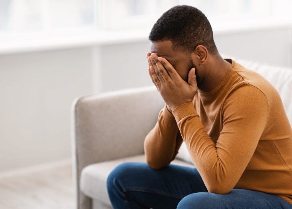 A depressed man wearing an orange shirt sits with his face in his hands while in a light-colored therapist's office