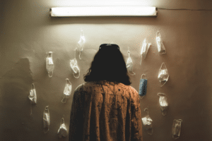 A woman stands in front of a dimly lit wall with many used medical masks on it. The woman is experiencing depression and anxiety after covid deemed post pandemic depression.